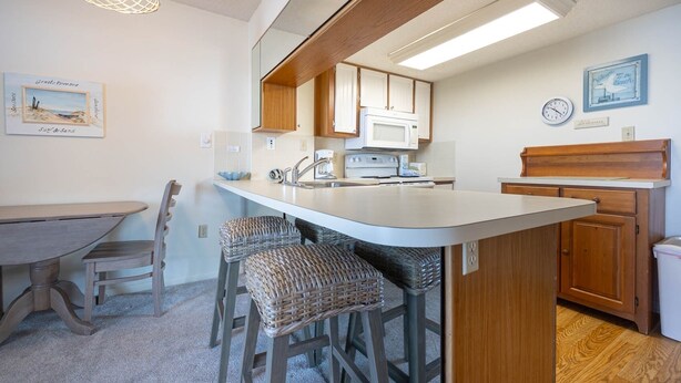 A compact kitchen with a central island, wooden cabinets, a microwave, and a sink. There are wicker bar stools and a dining table with chairs. - Gulf Shores Surf & Racquet 603a 1 Bedroom Condo (Gulf Shores)