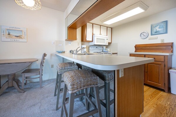 A compact kitchen with a central island, wooden cabinets, a microwave, and a sink. There are wicker bar stools and a dining table with chairs.