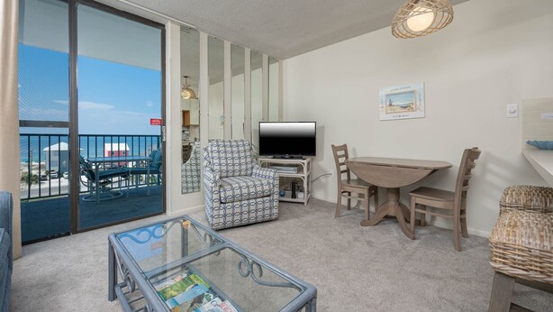 A living room with a glass coffee table, a TV, and a dining area with chairs. - Gulf Shores Surf & Racquet 603a 1 Bedroom Condo (Gulf Shores)