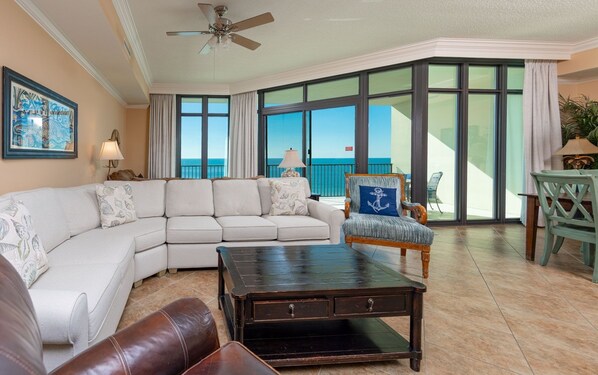 A living room with a large window offering a view of the ocean, a ceiling fan, and a dark wooden coffee table. - Phoenix West 1202 3 Bedroom Condo (Orange Beach)