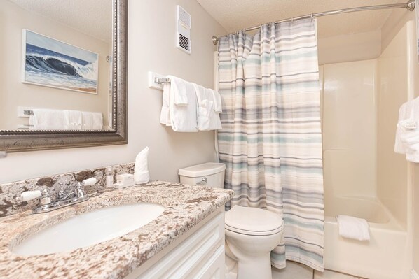 A bathroom with a marble sink, a toilet, a shower with a striped curtain, and a mirror with a painting of the ocean. - Island Winds West 2723-4 4 Bedroom Condo (Gulf Shores)