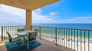 Ein Balkon mit Tisch und Stühlen mit Blick auf einen Strand mit blauen Sonnenschirmen und dem Meer.