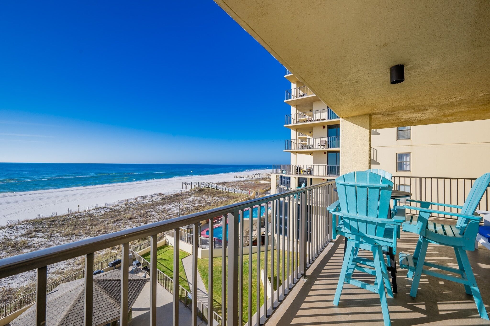 A balcony with a view of the beach, turquoise chairs, and a pool area.