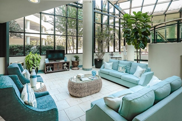 A spacious living area with a large window, a sofa set, a television, and various plants.
