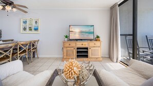 A living room with a TV, wooden cabinet, dining table, chairs, and a sofa. - Gulf Shores Surf & Racquet 610a 2 Bedroom Condo (Gulf Shores)