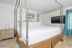 Una cama con dosel, ropa de cama blanca, una mesita de noche de madera, un televisor en la pared y un cuadro sobre la cama.