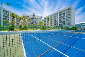 A tennis court with a net, surrounded by a fence, and adjacent to a multi-story building with balconies. - Gulf Shores Surf & Racquet 502b 2 Bedroom Condo (Gulf Shores)