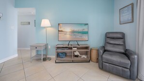 A living room with a flat-screen TV, a grey leather recliner, a small wooden side table, and a lamp.