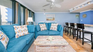 A room with a blue sofa set, a ceiling fan, a bar area with chairs, and a view through large windows. - Island Winds West 470 3 Bedroom Condo (Gulf Shores)