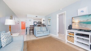 A living room with a sofa, TV stand, and a kitchen in the background. - Island Winds West 174 2 Bedroom Condo (Gulf Shores)