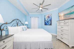 A bedroom with a bed, a ceiling fan, a dresser, a nightstand, a lamp, a painting, and a window with curtains. - Phoenix On The Bay 1223 2 Bedroom Condo (Orange Beach)