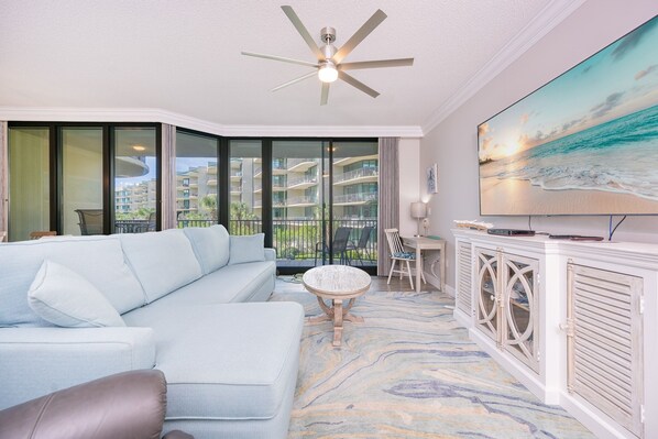 A modern living room with a light blue sofa, a round coffee table, and a television on a white cabinet. - Phoenix On The Bay 1223 2 Bedroom Condo (Orange Beach)