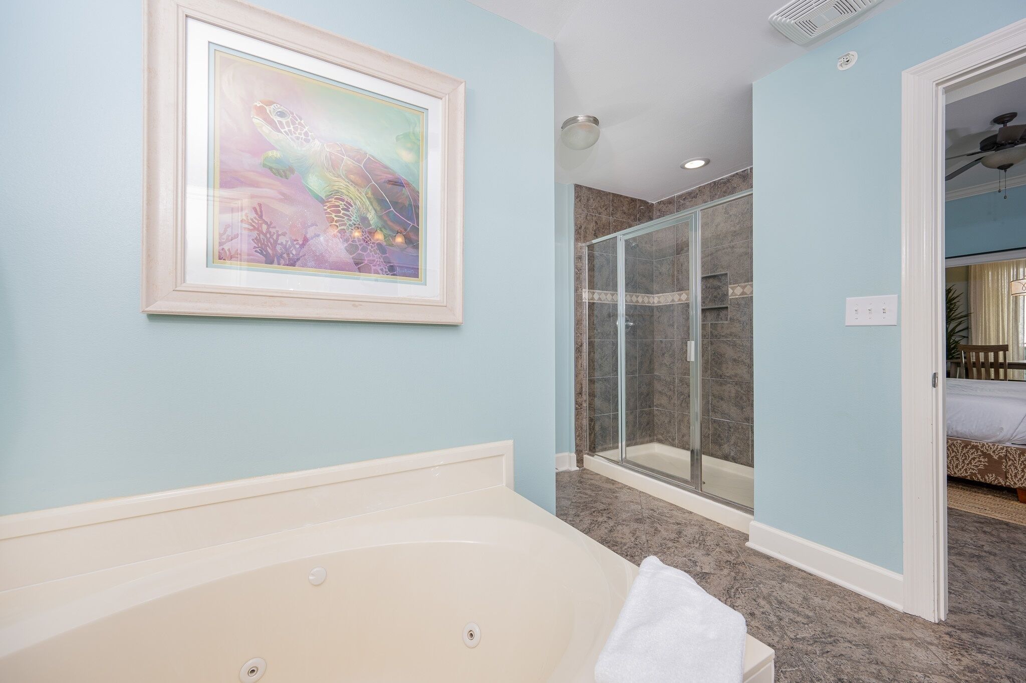 A bathroom with a bathtub, a framed artwork, a glass shower enclosure, and a visible bedroom with a bed.