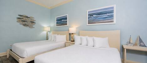 A room with two beds, a ceiling fan, wall art, and a small table with a model ship.