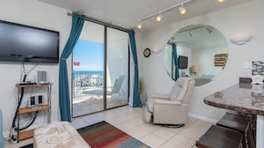 A living room with a TV, a chair, a small table, and a view through a sliding glass door. - Gulf Shores Surf & Racquet 605a 1 Bedroom Condo (Gulf Shores)