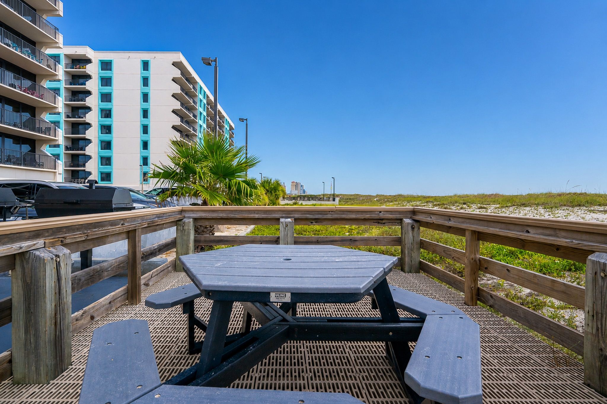 Une terrasse en bois avec une table de pique-nique et des bancs, d’où l’on a vue sur une plage et un immeuble doté de balcons.