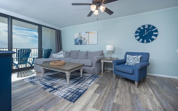 A modern living room with a grey sofa, a blue armchair, a wooden coffee table, and a large clock on the wall. - Island Winds West 872 2 Bedroom Condo (Gulf Shores)