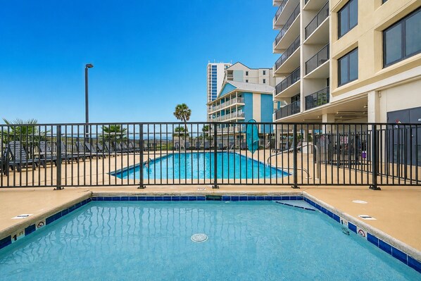 A swimming pool with a black metal fence, surrounded by a building with balconies and a clear blue sky. - Island Winds West 872 2 Bedroom Condo (Gulf Shores)