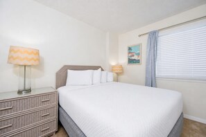 Ein ordentlich bezogenes Bett mit weißer Bettwäsche, ein Korbnachttisch mit Schubladen, eine Lampe mit Zickzackmuster und ein gerahmtes Bild an der Wand.