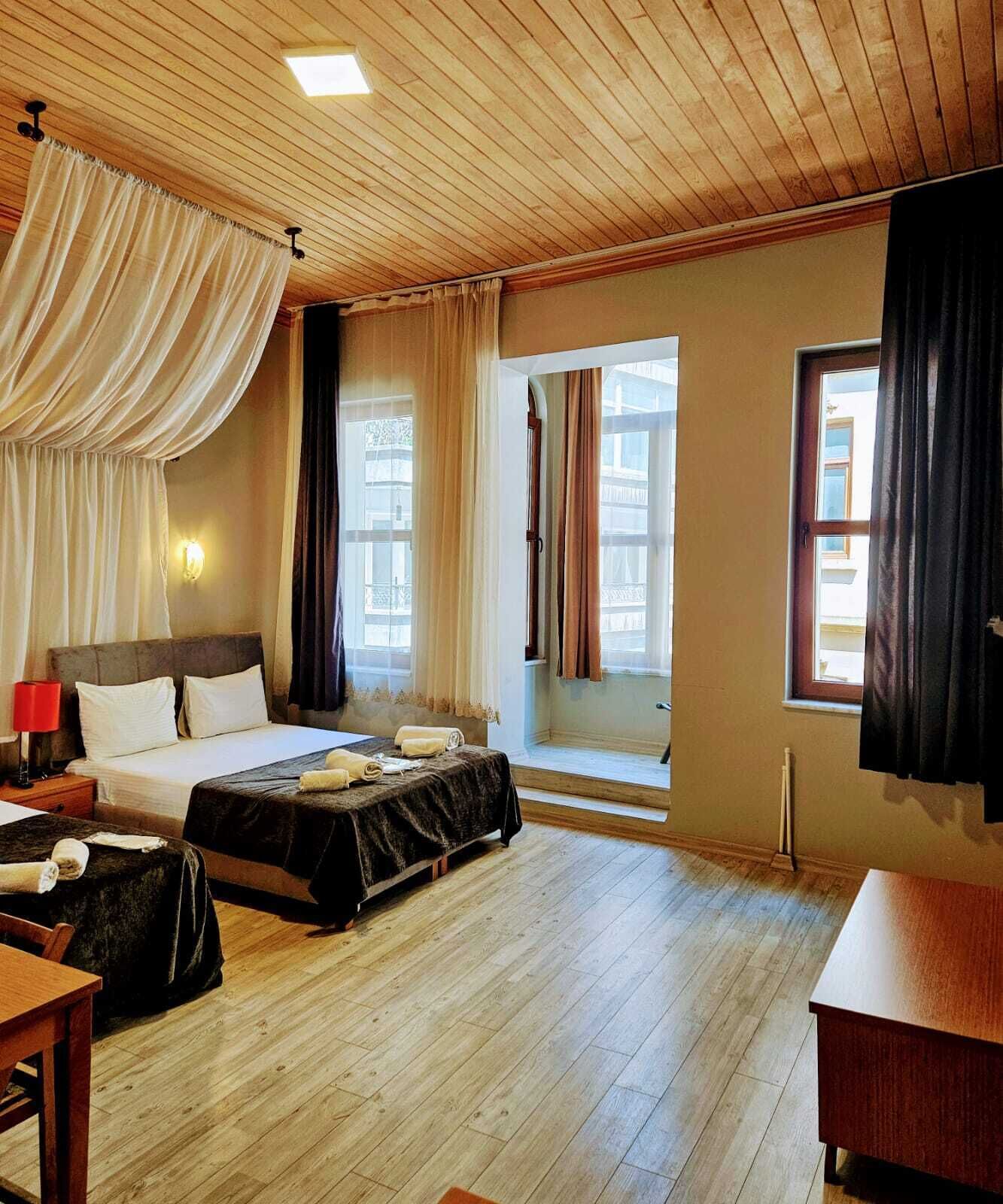 A hotel room with a wooden ceiling, two beds, a desk, and large windows with curtains.