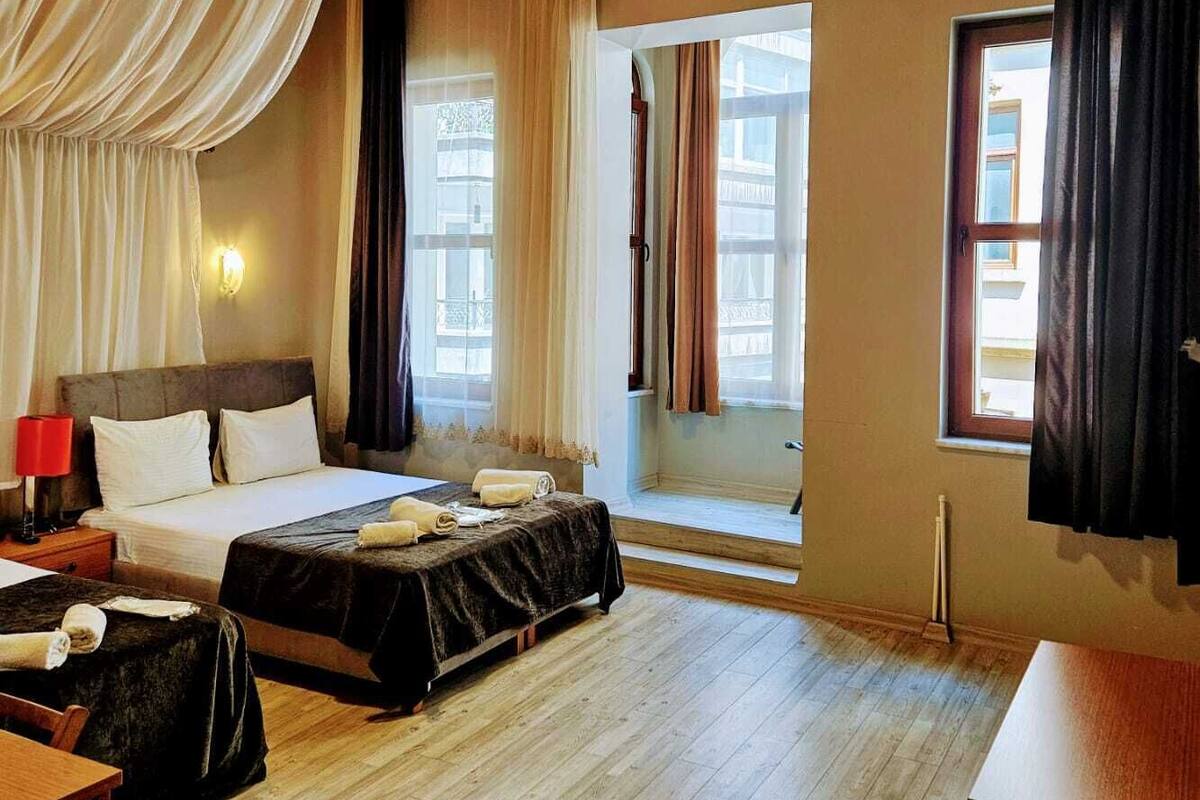 A hotel room with a wooden ceiling, two beds, a desk, and large windows with curtains.