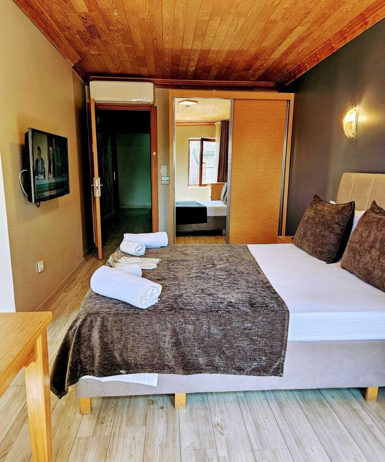 A bedroom with a bed, a wooden ceiling, a TV on the wall, and a bathroom visible through an open door.