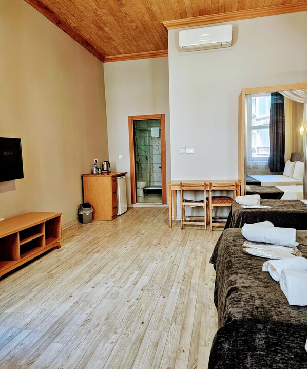 A hotel room with a wooden floor, a bed with a dark cover, a small table, a TV, and a shower area.