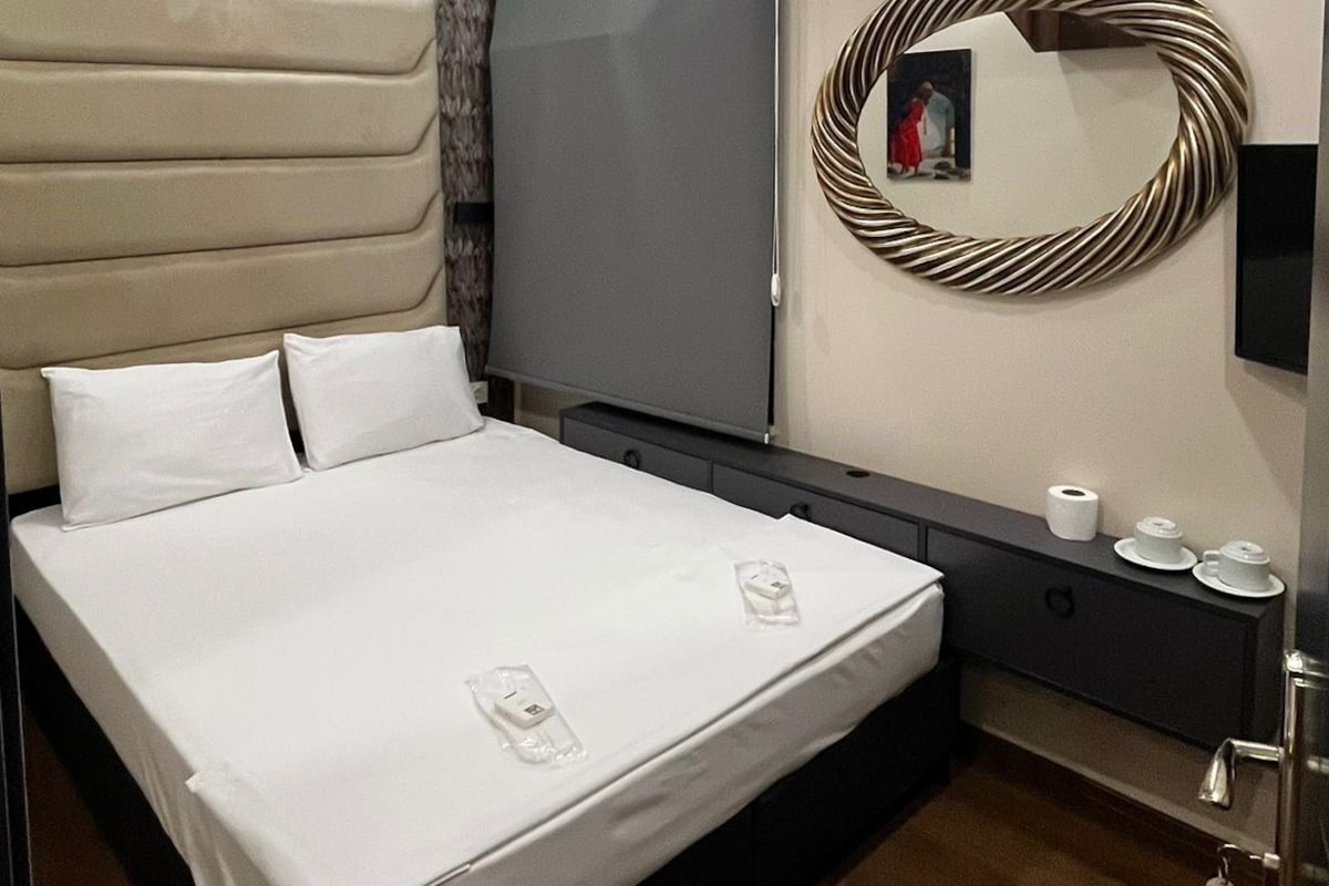 A modern bedroom with a large bed, a built-in headboard, a wall-mounted air conditioner, a mirror, and a bedside table with toiletries.