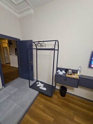A hotel room with a wooden floor, a desk with a coffee maker, a small trash bin, and a coat rack with shoes.