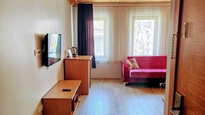 Una stanza con soffitto in legno, televisore a muro, una scrivania in legno, un divano rosso e una finestra con tende.