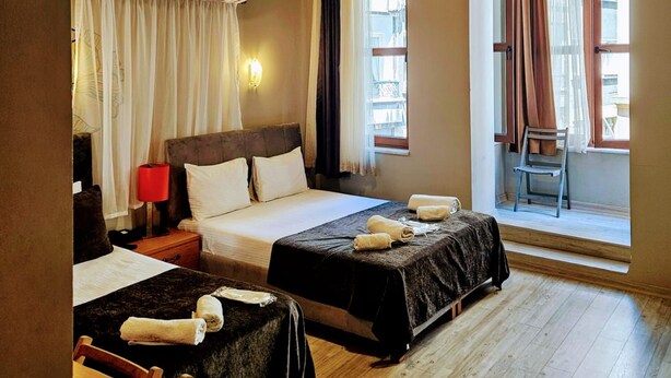 Una camera d'albergo con due letti, un soffitto in legno, una finestra con tende, una sedia e un comodino con una lampada.