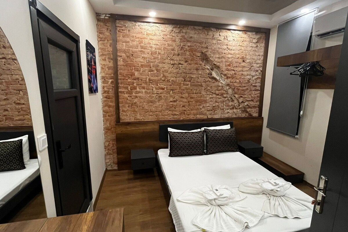 Una habitación con pared de ladrillo, cama con ropa blanca, cabecero de madera y una mesita pequeña.