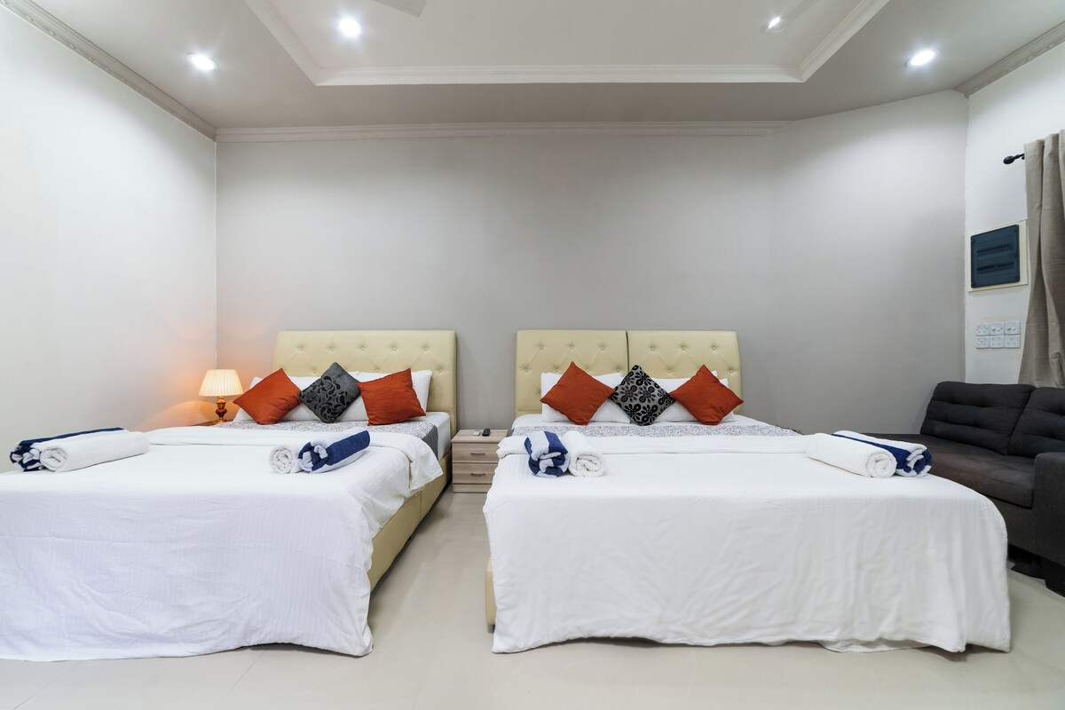 A hotel room with two beds, each with white bedding and decorative pillows. There is a sofa and a lamp on the left side.