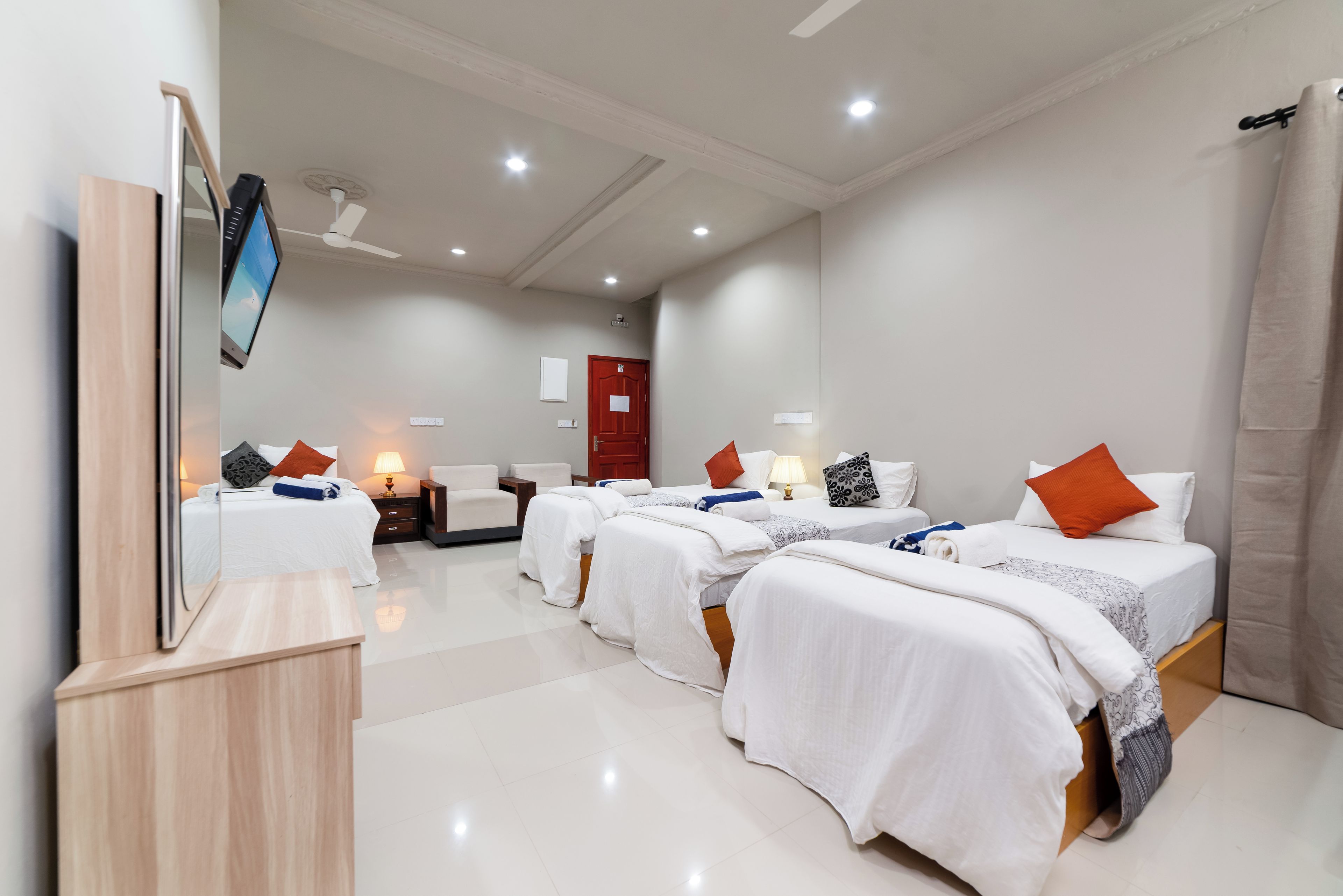 Ein Hotelzimmer mit drei Betten, einem Flachbildfernseher, einem Deckenventilator und einer roten Tür.