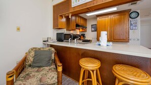 A compact kitchen with wooden cabinets, a microwave, a coffee maker, and a bar stool. - Gulf Shores Surf & Racquet 503a 1 Bedroom Condo (Gulf Shores)