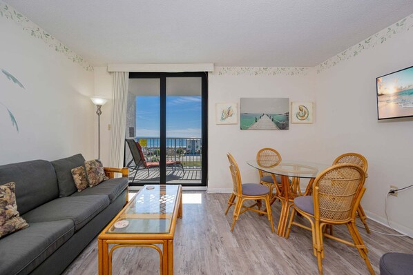 Una sala de estar con sofá, mesa de centro y sillones de mimbre. Hay un balcón con vistas a la ciudad y a la playa.