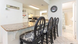 A modern kitchen with granite countertops, a dining area with black chairs, and a hallway leading to a bathroom. - Island Winds West 671 2 Bedroom Condo (Gulf Shores)