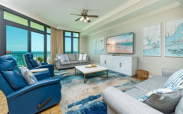 A spacious living room with a large window overlooking the ocean, a ceiling fan, a flat-screen TV, and nautical-themed wall art. - Phoenix West 305 3 Bedroom Condo (Orange Beach)