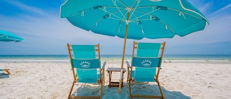 Two beach chairs with a table under a large umbrella on a sandy beach.