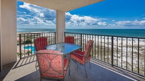 A balcony with a glass table and chairs, overlooking a beach and a swimming pool.