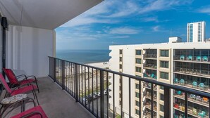 A balcony with red chairs, a small table, and a view of the ocean and a building with balconies.