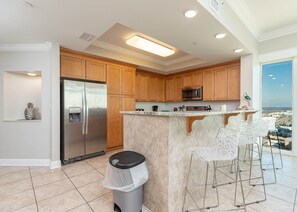 A modern kitchen with wooden cabinets, stainless steel appliances, a central island with bar stools, and a view through a large window. - Phoenix Gulf Shores 2004-1 1 Bedroom Condo (Gulf Shores)