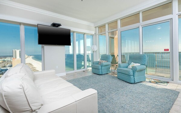 A spacious living room with a white sofa, two patterned armchairs, a flat-screen TV, and large floor-to-ceiling windows offering a view of the beach and cityscape. - Phoenix Gulf Shores 2004-1 1 Bedroom Condo (Gulf Shores)