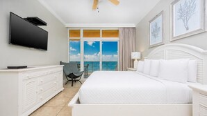 Ein Hotelzimmer mit einem Bett, einem Fernseher, einem Deckenventilator, einem Schminktischchen, einem Sessel und einem großen Fenster mit Blick auf den Himmel und die Wolken.