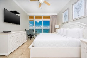 A hotel room with a bed, a TV, a ceiling fan, a dresser, a chair, and a large window with a view of the sky and clouds. - Phoenix Gulf Shores 2004-1 1 Bedroom Condo (Gulf Shores)