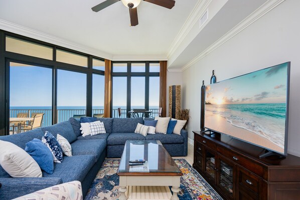 Ein Wohnzimmer mit großem Fernseher, einer Couch und einem Couchtisch. Ein Balkon bietet Blick auf den Ozean.