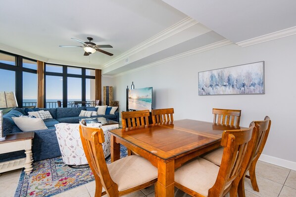 A living room with a wooden dining table, blue sofa, and a TV on the wall. - The Oasis 2905 Formerly Phoenix West Ii 2905 3 Bedroom Condo (Orange Beach)