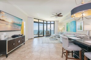 A modern hotel room with a large window, a ceiling fan, a dining area, and a view of the ocean. - The Oasis 2103 Formerly Phoenix West Ii 2103 3 Bedroom Condo (Orange Beach)
