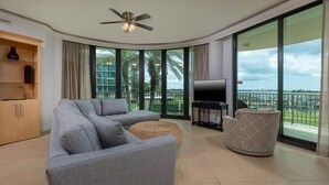 A modern living room with a grey sectional sofa, a round ottoman, a flat-screen TV on a stand, and a ceiling fan. - Phoenix On The Bay 1103 4 Bedroom Condo (Orange Beach)