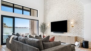 Una sala de estar moderna con un sofá seccional gris, un televisor de pantalla plana montado en una pared de ladrillo, y un ventanal con vistas a edificios.